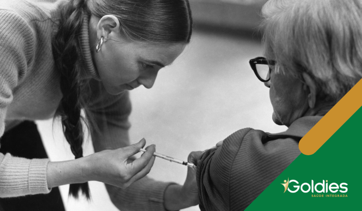 Um profissional de saúde administra uma vacina a um idoso nesta imagem em preto e branco. O logotipo da Goldies Saúde Integrada é visível no canto.