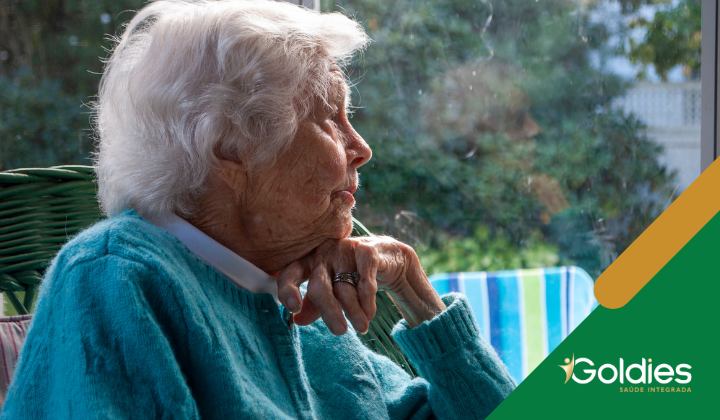 Mulher idosa com cabelos brancos e suéter azul olha pensativamente para fora de uma janela; a luz do sol ilumina suavemente seu rosto. Logotipo da Goldies Saúde Integrada no canto.