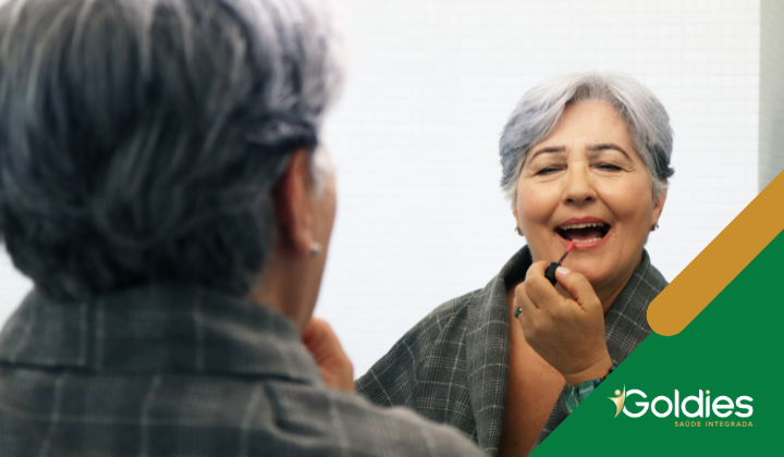 Pessoa idosa com cabelo curto e grisalho aplica batom, sorrindo para seu reflexo no espelho. A pessoa está usando um robe cinza. O logotipo no canto diz Goldies Saúde Integrada.