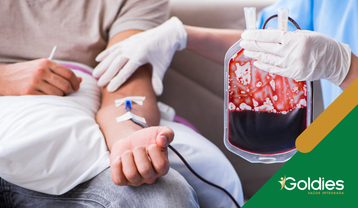 Pessoa doando sangue, com um profissional de saúde usando luvas segurando uma bolsa de sangue cheia e o braço de uma pessoa conectado a uma linha intravenosa. O logotipo no canto diz Goldies Saúde Integrada.