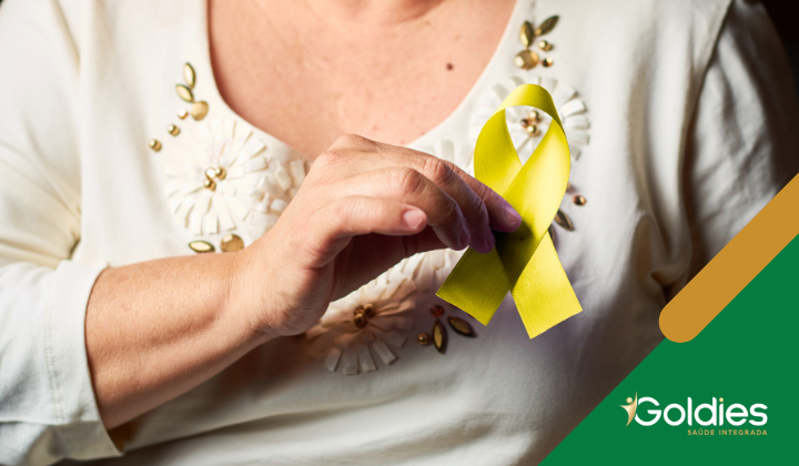 Pessoa em uma camisa branca com detalhes florais segurando uma fita amarela. O logotipo do Goldies está no canto inferior direito.