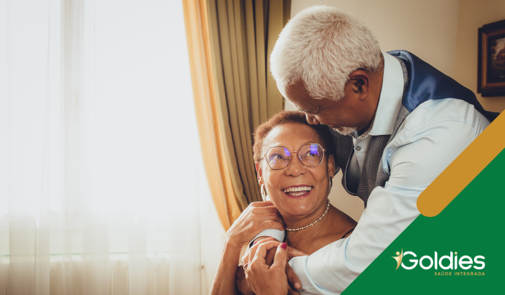 Homem idoso abraça uma mulher sorridente em uma sala bem iluminada. Ambos estão vestidos formalmente. Um logotipo Goldies está no canto inferior direito.