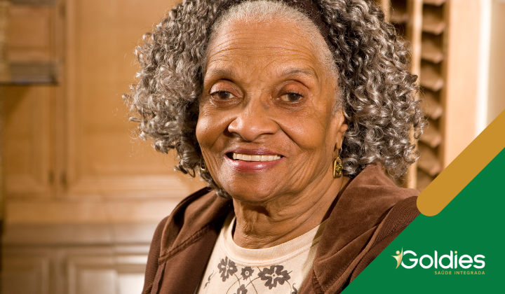 Mulher idosa com cabelos grisalhos cacheados sorrindo calorosamente, vestindo uma jaqueta marrom e blusa estampada, dentro de casa. O logotipo da Goldies é visível no canto.