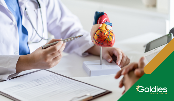 Um médico explicando um modelo de coração para um paciente, com uma prancheta e caneta na mesa. O logotipo Goldies é visível no canto.