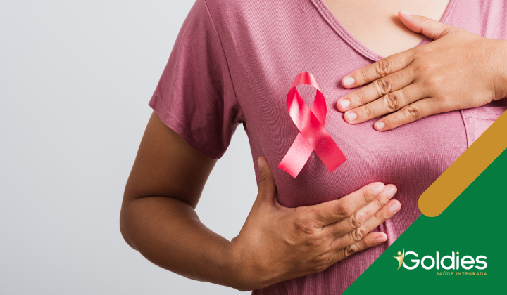 Uma pessoa vestindo uma camisa rosa segura uma fita rosa no peito, simbolizando a conscientização sobre o câncer de mama. O canto inferior direito tem um logotipo com o nome Goldies e um slogan.