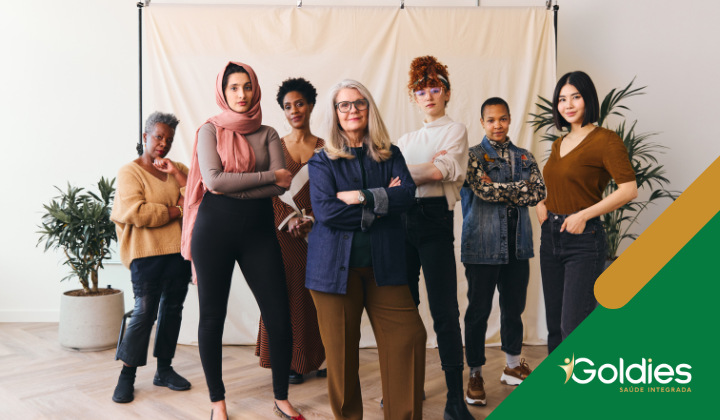 Um grupo diverso de sete mulheres em pé confiantemente em frente a um cenário bege, com duas plantas de cada lado. Um logotipo que diz Goldies Saúde Integrada está no canto.