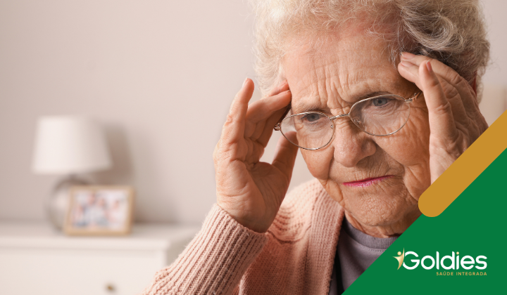 Mulher idosa com óculos segurando as têmporas, parecendo ter dor de cabeça, em uma sala suavemente iluminada. Uma luminária e uma moldura de foto estão desfocadas no fundo. O logotipo diz Goldies.