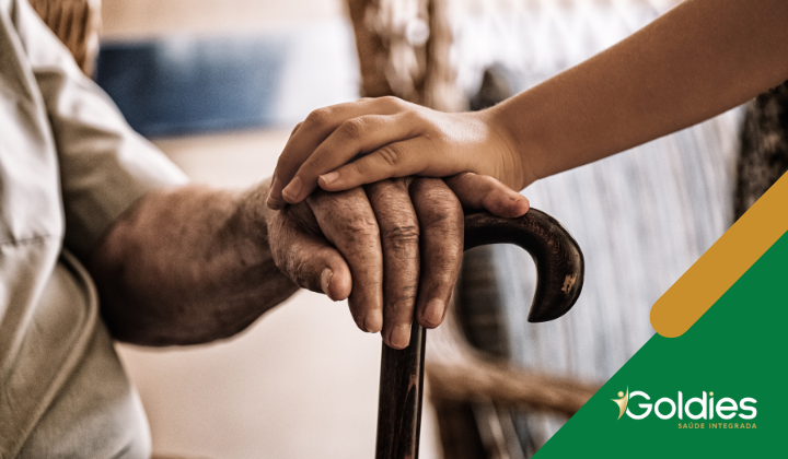 Uma mão jovem repousa gentilmente sobre a mão de uma pessoa idosa segurando uma bengala, simbolizando cuidado e apoio. O logotipo da Goldies Saúde Integrada é visível no canto.