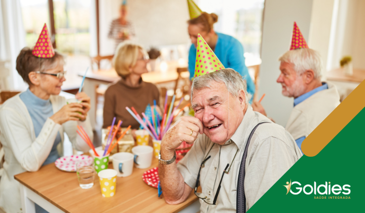 Pessoas idosas usando chapéus de festa, sorrindo, sentadas ao redor de uma mesa com copos coloridos, canudos e bebidas. Atmosfera alegre e brilhante.