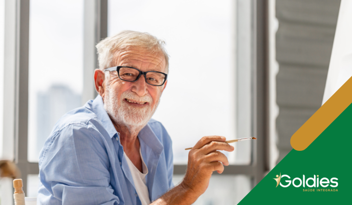 Homem idoso com óculos e barba, segurando um pincel, sorrindo para a câmera. Ele está sentado em uma sala iluminada. O logotipo Goldies Saúde Integrada é visível no canto.