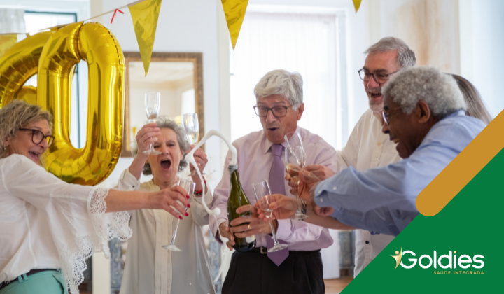 Um grupo de idosos comemorando com champanhe em ambientes fechados, segurando taças sob números de balões dourados. Eles estão sorrindo e comemorando.