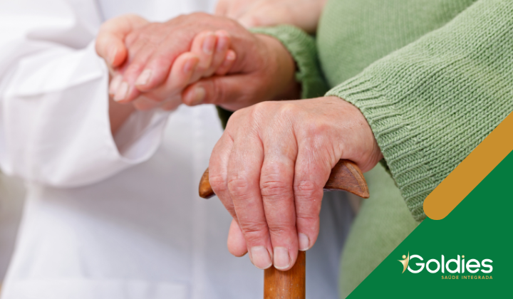 Close de uma pessoa idosa segurando uma bengala de madeira, com um profissional de saúde segurando gentilmente sua mão. Logotipo da Goldies Saúde Integrada no canto inferior direito.