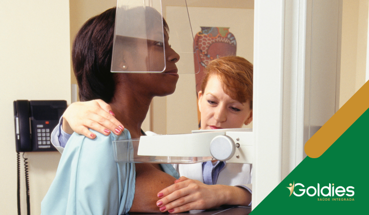 Um profissional de saúde auxilia uma mulher que está passando por uma mamografia. O cenário é um consultório médico. O logotipo Goldies Saúde Integrada é visível no canto.