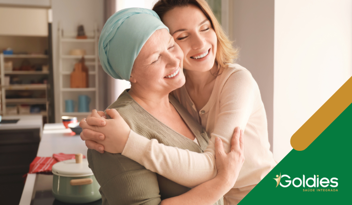 Duas mulheres se abraçando em uma cozinha. Uma está usando um lenço na cabeça, sorrindo com os olhos fechados. Um logotipo com Goldies Saúde Integrada está no canto inferior direito.